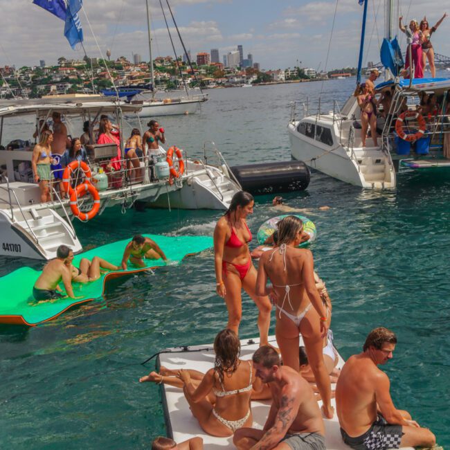 People in swimsuits relax and socialize on a floating platform and nearby boats in a lively harbor, with city buildings and greenery in the background under a partly cloudy sky. "Yacht Social Club" logo is visible in the corner.