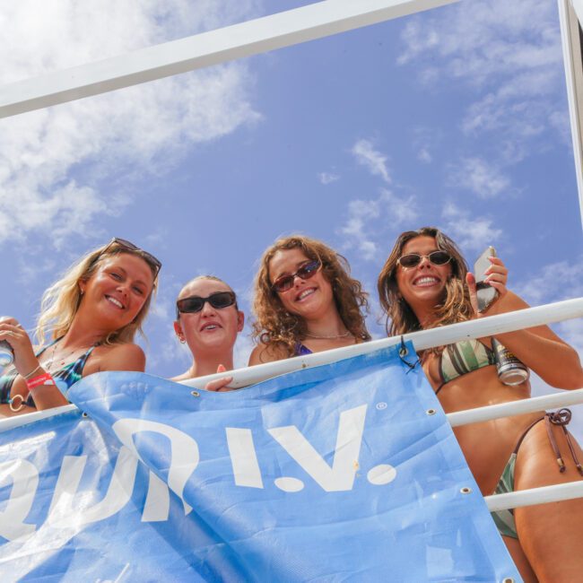 Four women in swimsuits smile and pose for a photo from behind a blue railing under a sunny sky with scattered clouds. They each hold drinks and wear sunglasses, enjoying an outdoor event.