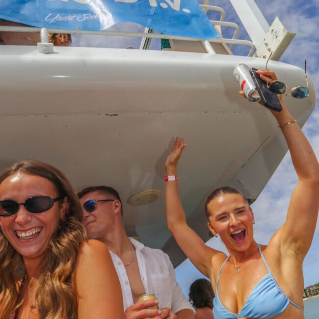 Three young adults enjoy a sunny day on a boat; one woman in a blue bikini raises her arms and smiles, while another woman and a man in sunglasses laugh beside her. Blue sky and clouds are visible above.