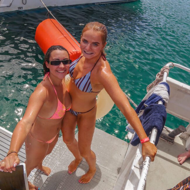 Two women in bikinis smile at the camera on the steps of a boat, with clear blue water in the background. One wears sunglasses, and a boat with people is visible behind them. Both appear to be wet from swimming.