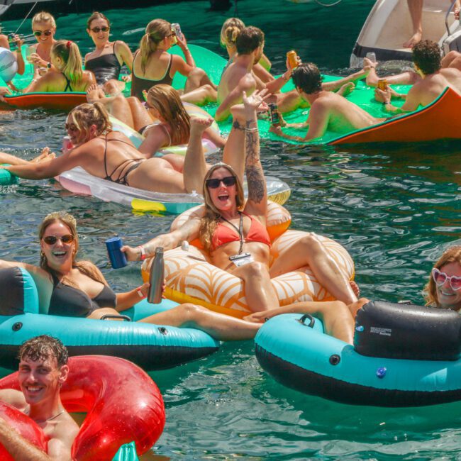 A group of people in swimsuits relax and have fun on colorful inflatable floats in the water, holding drinks and smiling on a sunny day. The scene is lively and festive, with boats and more people in the background.