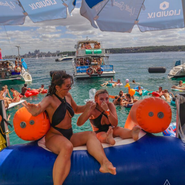 Two women in black swimsuits sit on an inflatable float under umbrellas on a boat, surrounded by people swimming and relaxing on floats, with boats and city skyline visible in the background.