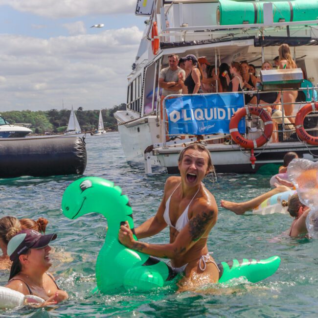 A woman in a swimsuit laughs while riding a green inflatable dinosaur in the water, surrounded by others. In the background, people are partying on a yacht under partly cloudy skies. "Yacht Social" is written on the image.