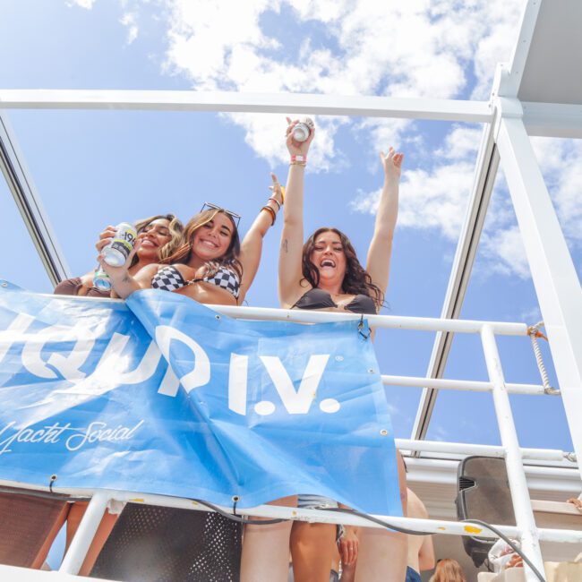 Three women in swimsuits stand on a deck, smiling and holding drinks, with one raising her arm in excitement. They are behind a blue banner under a bright, partly cloudy sky.