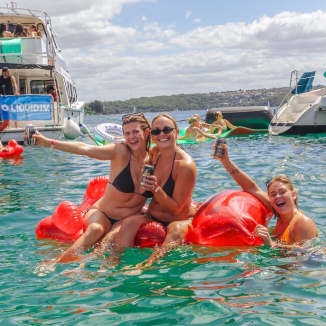 Three women in swimsuits sit on a red inflatable in the water, smiling and holding drinks. Other people are swimming or on floats nearby, with party boats and blue sky in the background.