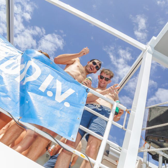 Two men in swimwear smile and give a thumbs-up on a boat deck, standing behind a blue "LIQUID I.V." banner. The sky is bright and partly cloudy, and other people are visible around them.