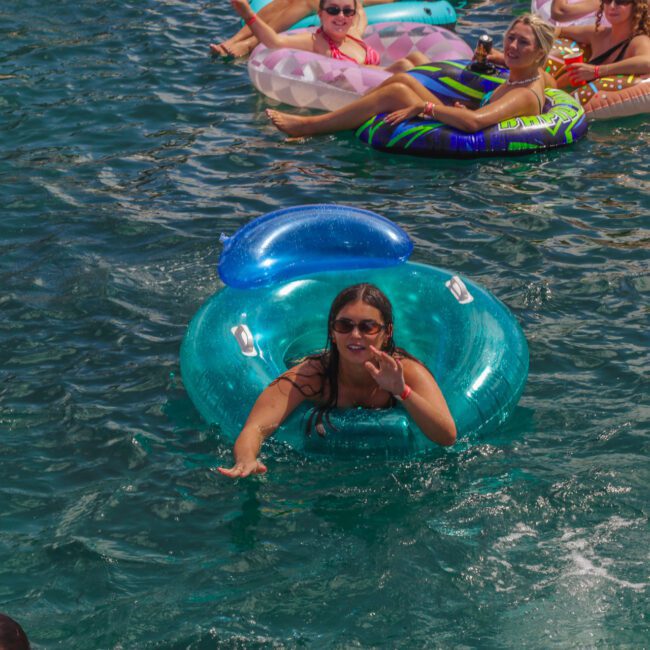 A woman in sunglasses floats on a blue inflatable ring in a sunny body of water, surrounded by other people relaxing on colorful pool floats.