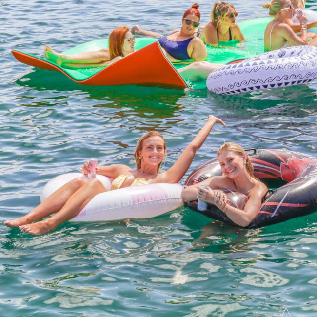 A group of women relax on colorful inflatables and floaties in clear blue water, smiling and enjoying sunny weather. Two women in the foreground pose and wave at the camera, holding drinks.