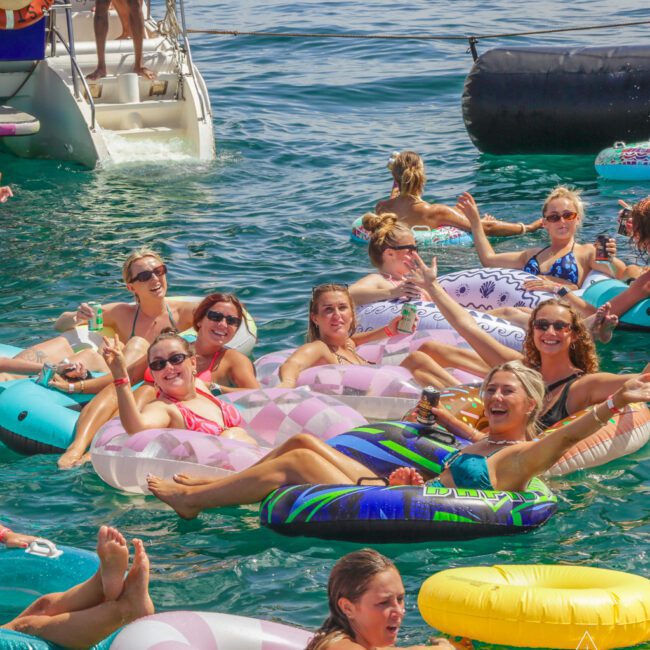 A group of women relax and smile on colorful inflatable rings in clear blue water near a docked boat, enjoying a sunny day together. Some wave and raise their drinks, creating a lively, festive atmosphere.