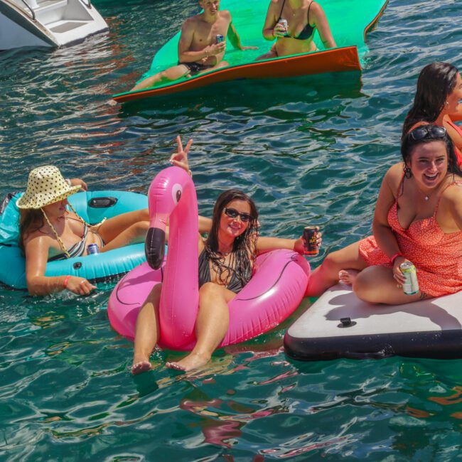 People relax on colorful inflatables in the water. A woman on a pink flamingo float smiles and flashes a peace sign, while others lounge nearby, enjoying drinks and the sunny weather.