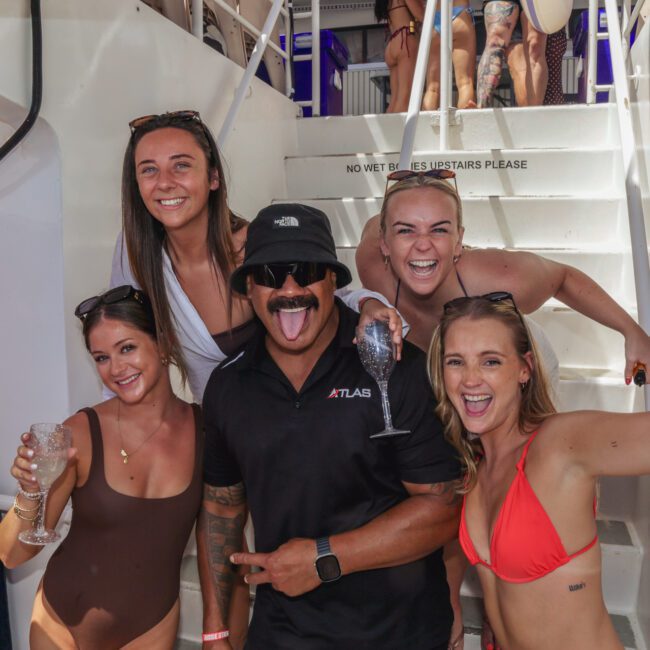 A group of five smiling adults in swimwear and sunglasses pose together on a boat, holding drinks, with white stairs and more people in the background. The atmosphere is festive and sunny.