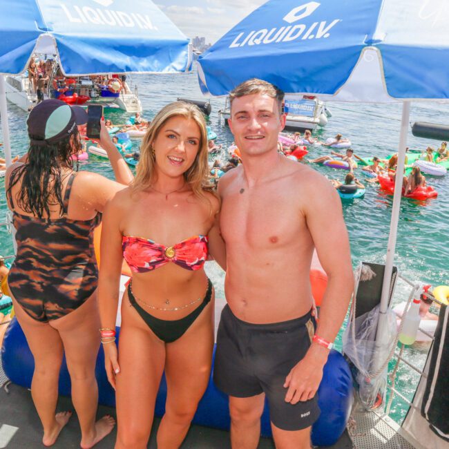 A woman and a man in swimsuits smile at the camera under blue umbrellas at a lively pool party on the water, surrounded by people floating on inflatables and enjoying the sunny day.