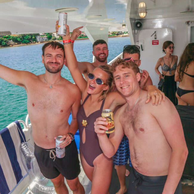 A group of friends in swimsuits smile and pose with drinks on a boat, with blue water and greenery in the background. Some are wearing sunglasses and wristbands, enjoying a sunny day.