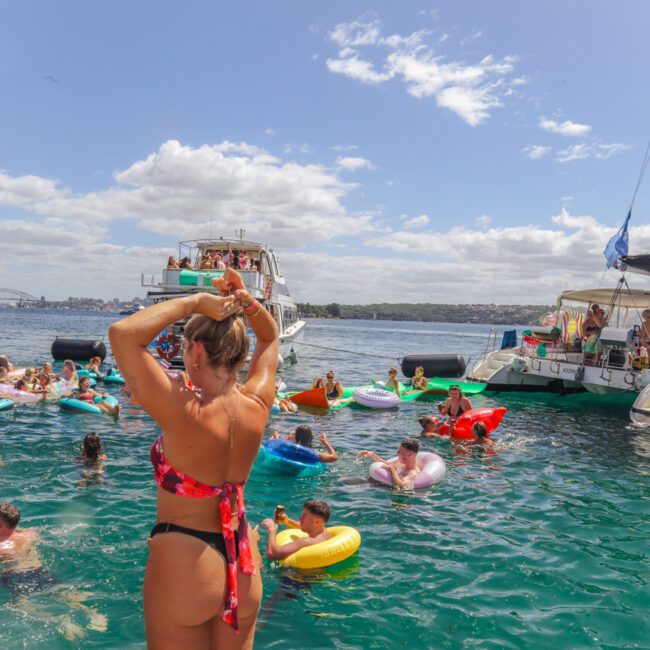 A woman in a pink bikini stands on a boat platform, facing a lively group of people relaxing on floaties and swimming near yachts on bright blue water under a partly cloudy sky.