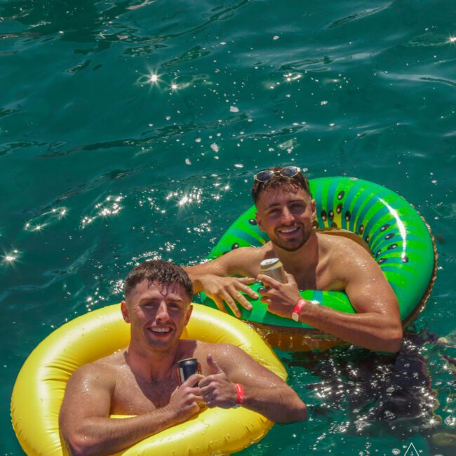Two smiling men in bright inflatable rings float on clear blue water, holding drinks and giving a thumbs-up. Sunlight sparkles on the water's surface.