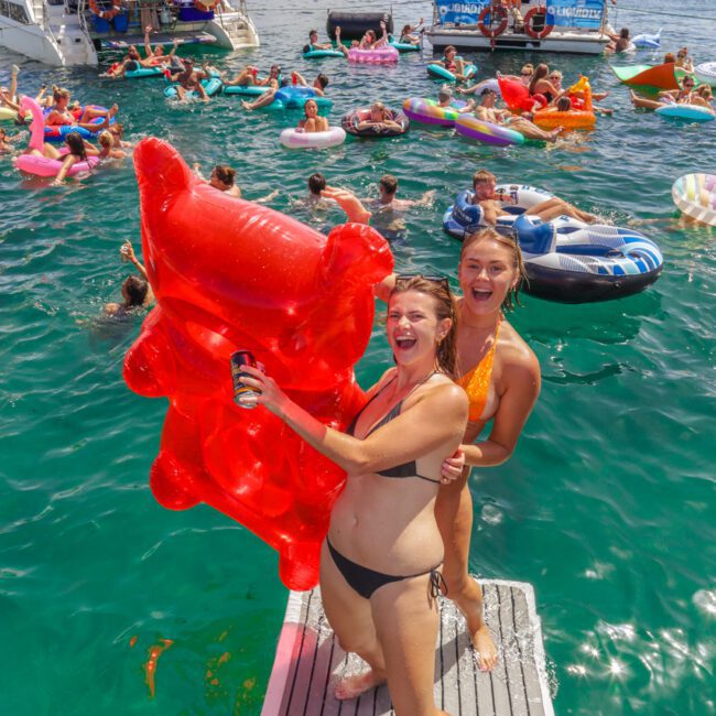 Two women in swimsuits laugh and pose with a large red inflatable on a boat’s platform, surrounded by people on floaties enjoying a lively party in turquoise water near anchored boats.