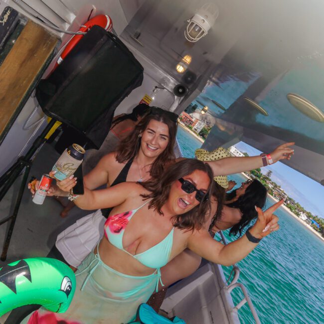 Two women in swimsuits smile and pose for the camera on a boat deck, holding drinks. They’re surrounded by inflatable pool toys, with the ocean and shoreline visible in the background. Other people are enjoying the party.