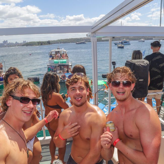A group of young adults in swimsuits smile and pose on a boat under sunny skies. The background shows other boats on the water and people enjoying a lively outdoor party.