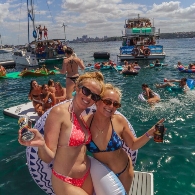 Two women in swimsuits and sunglasses smile and pose with drinks on a boat dock, surrounded by people on floats and boats enjoying a sunny day on the water. The background features boats, city skyline, and blue sky.