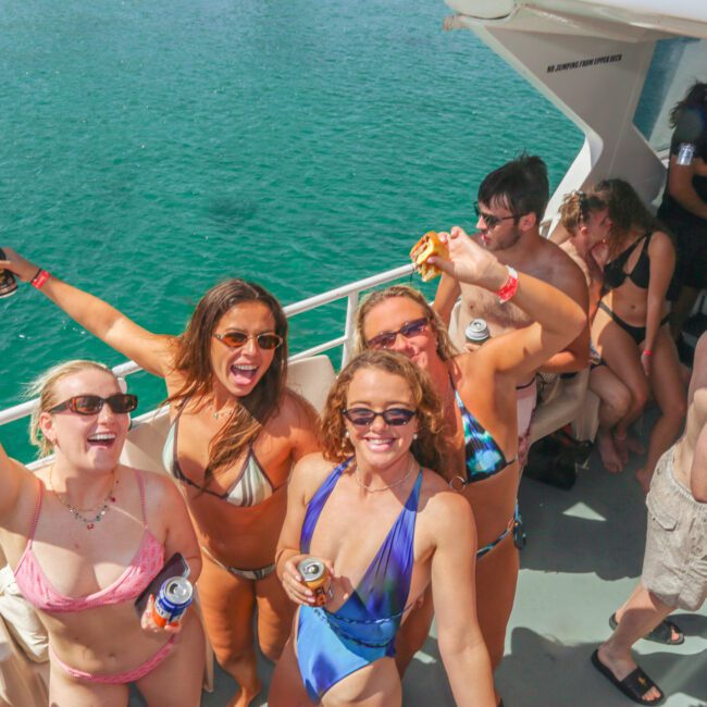 A group of young adults in swimsuits enjoy a lively party on a boat, smiling, holding drinks, and taking selfies under the sunny sky with bright blue water in the background.