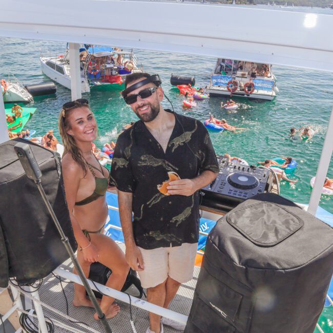 A smiling woman in a bikini and a man in sunglasses and a hat stand beside DJ equipment on a boat, surrounded by speakers, with people floating and boats on the water in the background.