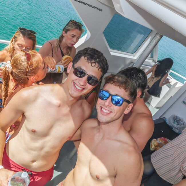 A group of young adults in swimsuits enjoy a sunny day on a boat, with two men in the foreground smiling at the camera while wearing sunglasses, surrounded by friends and the ocean in the background.