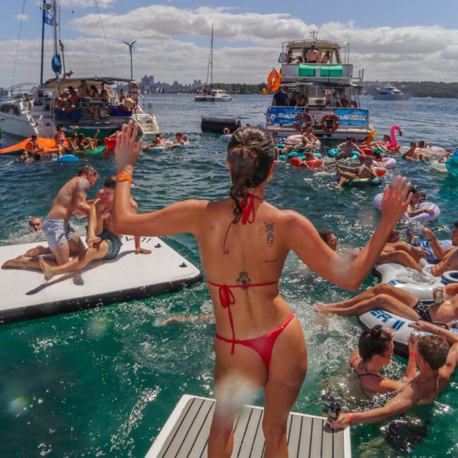 A woman in a red bikini stands on a boat platform, arms raised, facing people swimming and relaxing on floating mats in a lively, sunny harbor with boats and yachts in the background.