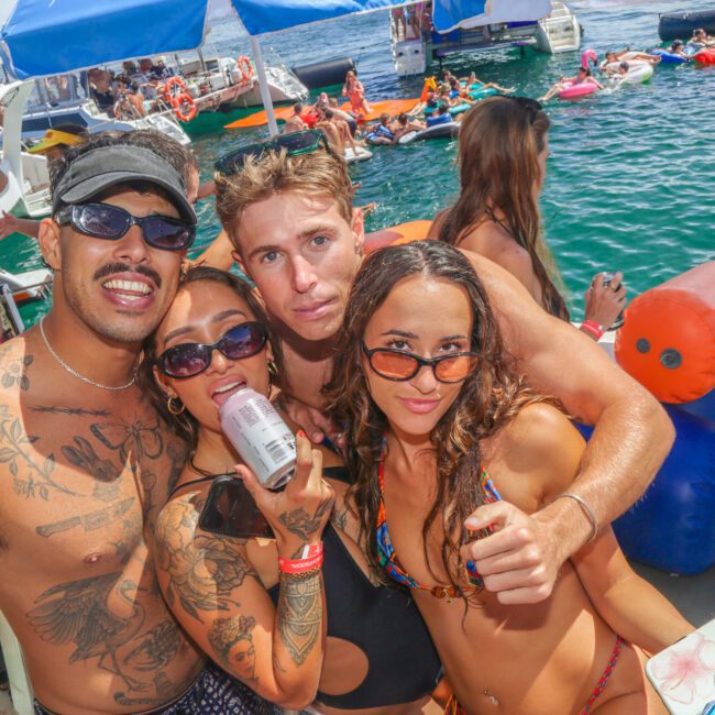 Four young adults in swimwear pose together on a crowded boat party, smiling at the camera. The background shows people swimming, lounging on inflatables, and enjoying a sunny day on the water.