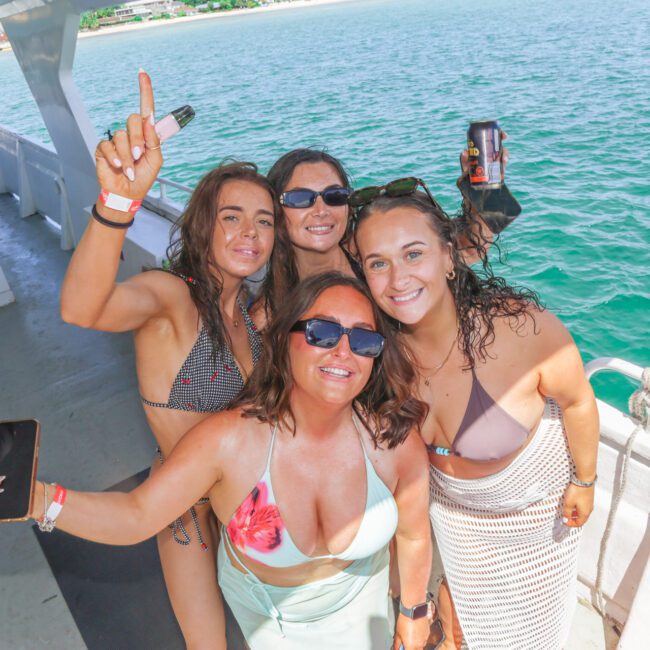 Four women in swimsuits and sunglasses pose and smile on a boat with drinks in hand, with turquoise water and a distant shoreline visible in the background on a sunny day.