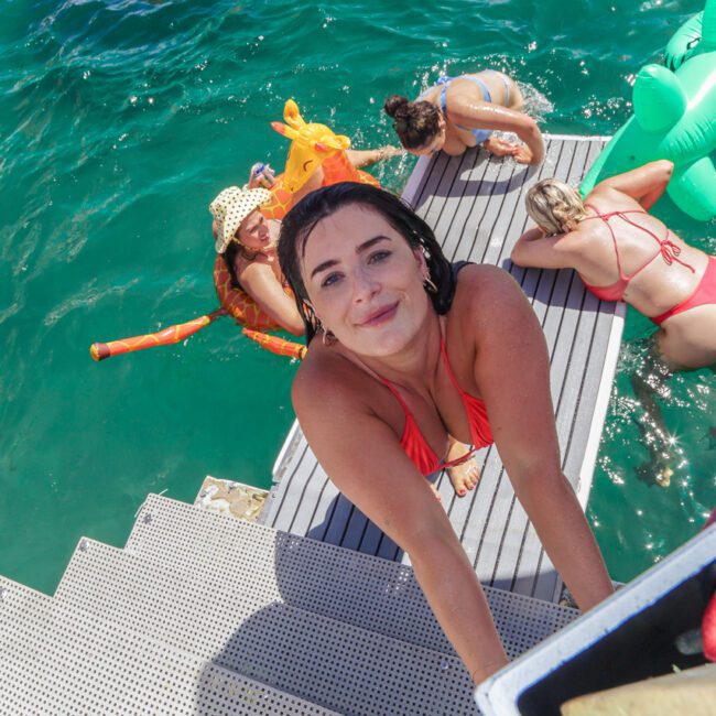 A woman in a red swimsuit climbs up a metal ladder from clear blue water, smiling at the camera. Other women in swimsuits relax and play with inflatables in the water behind her.