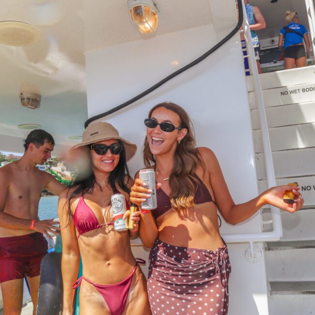 Two women in swimsuits and sunglasses smile and pose with drinks on a boat. Other people are in the background near stairs with a "No Wet Bodies Upstairs Please" sign. It’s sunny and the atmosphere is festive.