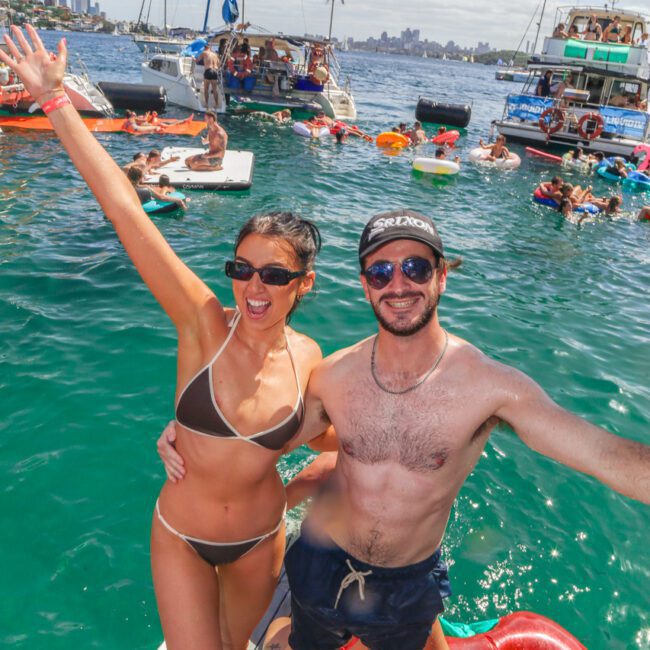 A smiling woman in a bikini and a man in swim trunks and sunglasses pose on a dock with boats and people swimming and relaxing on floaties in the background. It’s a sunny day on the water at a lively social event.