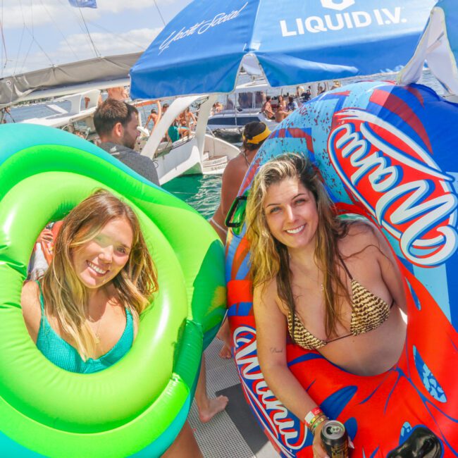 Two smiling women in swimsuits hold large inflatable pool floats—one green, one blue with red design—on a boat party. Other people and boats are visible on the water in the sunny background.