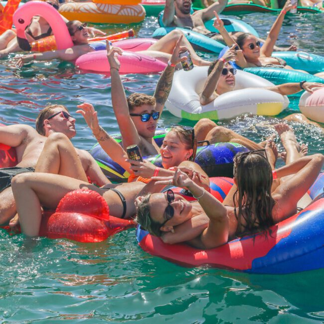 A group of people relax and have fun on colorful pool floats in the water near a boat, smiling, taking selfies, and enjoying drinks under the sun.