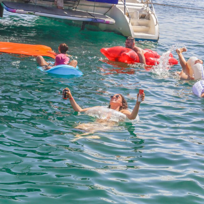 A group of people relax in the water near a yacht, some on inflatable floats. One person floats on their back, holding drinks, enjoying the sunny day. City buildings are visible in the background.
