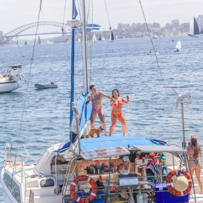 Two people in swimsuits dance on the roof of a boat with others below them, surrounded by sailboats on the water. The Sydney Harbour Bridge and city skyline are visible in the background under a partly cloudy sky.
