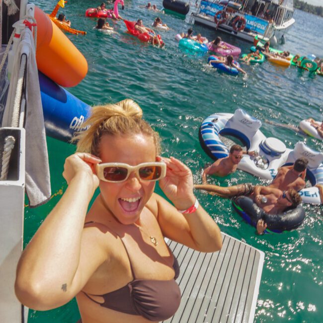 A woman in a brown bikini and sunglasses smiles at the camera on a dock, with boats and people floating on colorful inflatables in the water behind her on a sunny day.