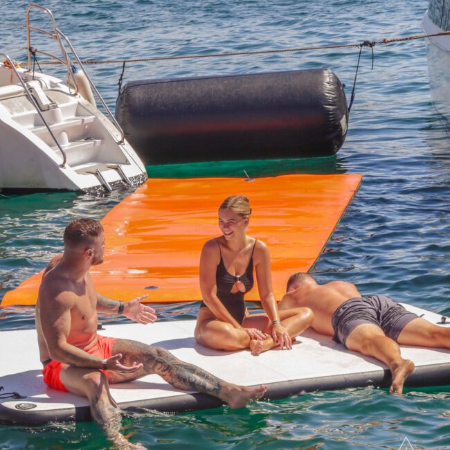 Three people relax on a floating mat by a boat in sunny weather; one man sits talking, a woman in a black swimsuit sits smiling, and another man lies down resting. The sea and an orange mat are visible in the background.