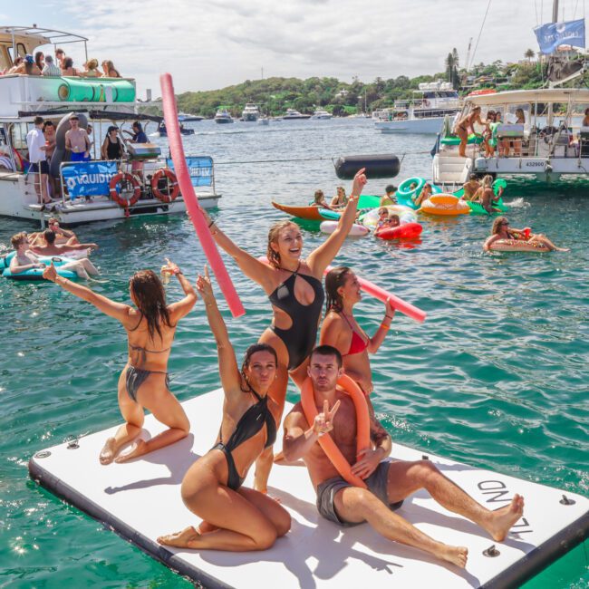 A group of people in swimsuits pose and have fun on a floating mat in the water, surrounded by others on inflatables and boats under a sunny sky during a lively summer party.