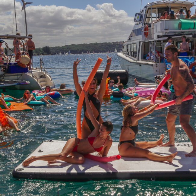 People in swimsuits play with pool noodles on a floating platform in the water, surrounded by others lounging on inflatables and boats. The atmosphere is festive and lively under a partly cloudy sky.