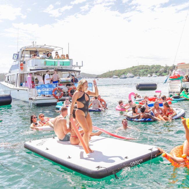 People relax and have fun on inflatable floats and paddleboards in the water near boats on a sunny day. A woman in a black bikini stands posing on a floating mat, while others socialize and enjoy the lively scene around her.