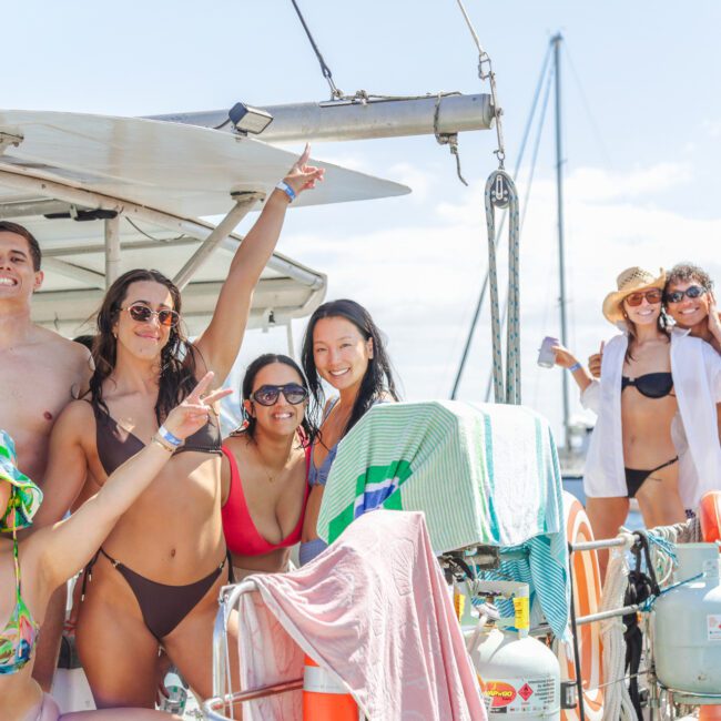 A group of smiling people in swimsuits pose and make peace signs on a sailboat, enjoying a sunny day on the water. Towels, hats, and boating equipment are visible, with a bridge and blue sky in the background.