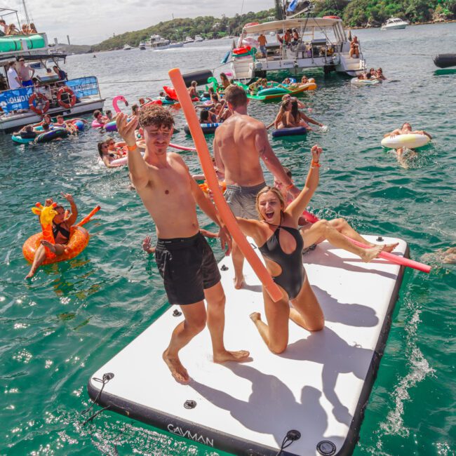 Two people on a floating platform, smiling and holding pool noodles, surrounded by others on inflatables and boats in a lively, sunny water party scene.