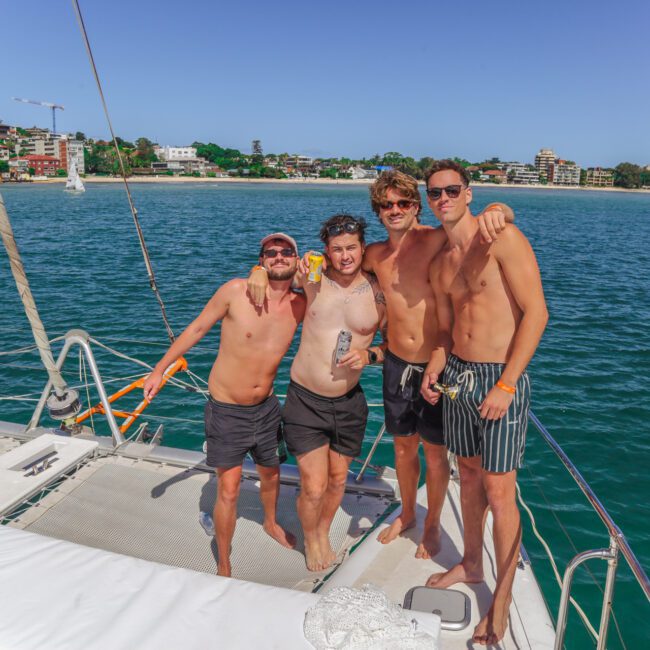 Four men in swim trunks stand together smiling on the deck of a yacht with water and coastal buildings in the background on a sunny day.