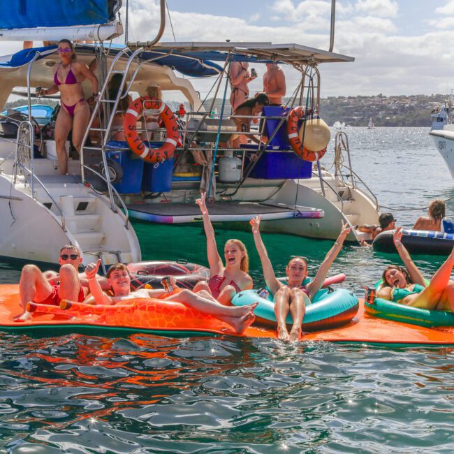 A group of people relax on colorful floaties in the water near a docked boat, enjoying a sunny day. Other people are on the boat, and the background shows a shoreline and partly cloudy sky.