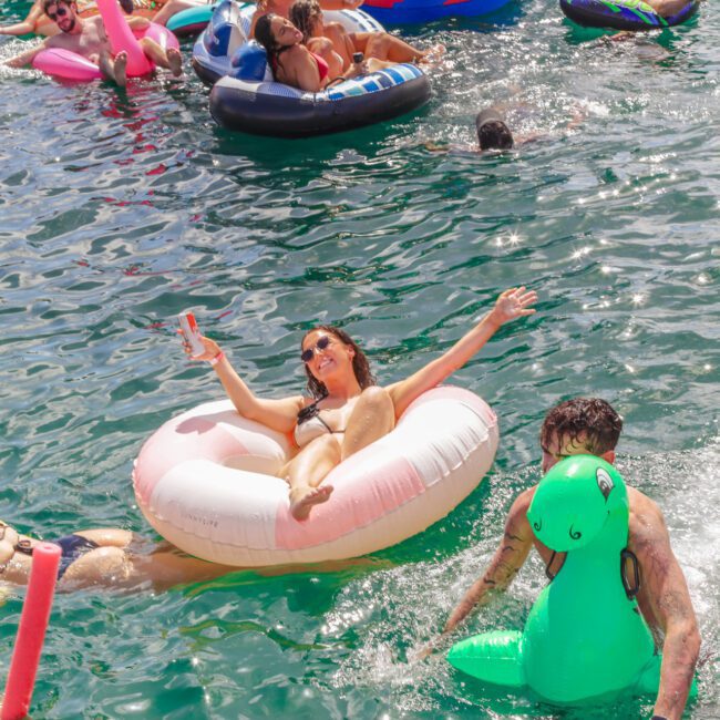 People relax and have fun on colorful pool floats in a sunny body of water. A smiling woman lounges on a donut float, taking a selfie, while others swim or rest on inflatables around her.