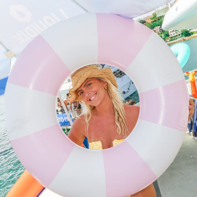 A woman in a straw hat and yellow bikini smiles while posing behind a large pink and white inflatable swim ring on a boat by the water.