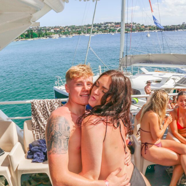 A young couple embraces and smiles on the deck of a boat, surrounded by friends in swimwear. The boat is on the water near a coastline with houses and greenery visible in the background.