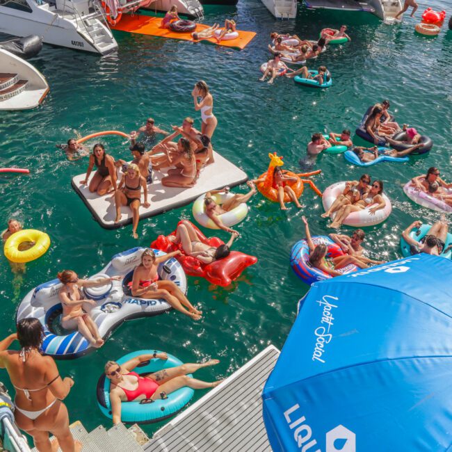 A group of people relax on colorful inflatables in the water between yachts at a lively pool party. Some stand on a floating platform, while others float nearby. A large blue umbrella is in the foreground.