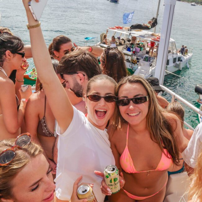 A group of young people in swimsuits and sunglasses smile and hold drinks while enjoying a lively boat party under sunny skies, with other boats and a calm sea in the background.